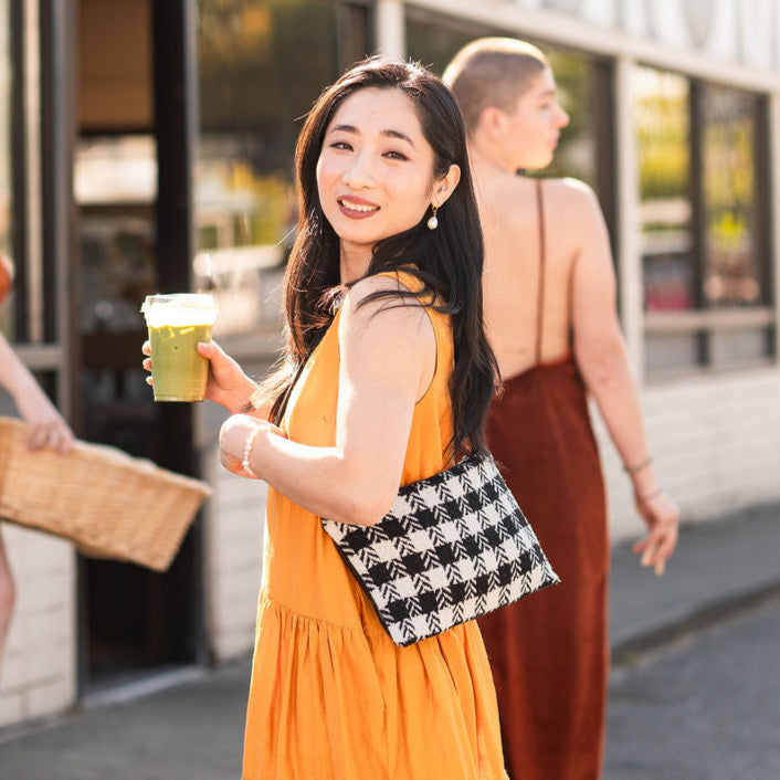 Woman in a yellow dress holding a green cup wearing a houndstooth bag outside a store with other people in the background.
