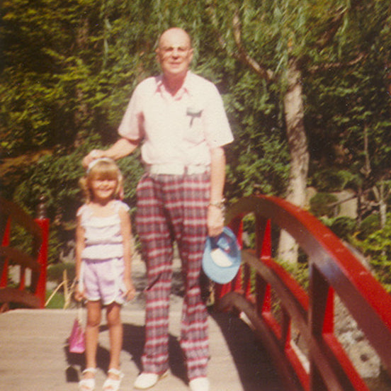 grandpa in plaid pants and young girl holding a pink purse, standing on a red bridge with greenery in the background at Manito Park, Spokane WA