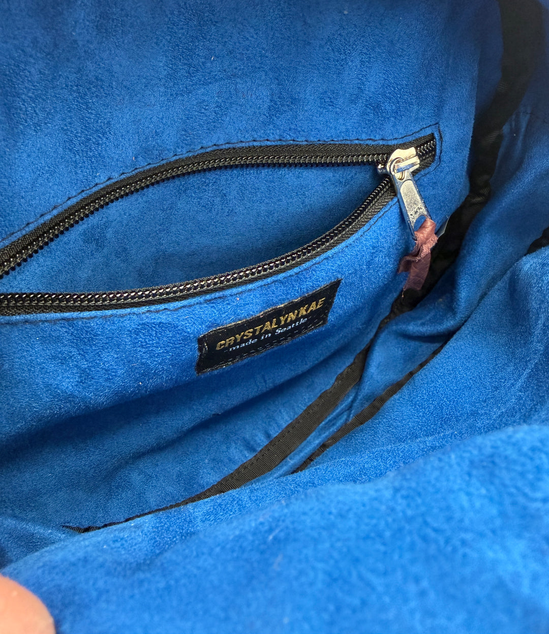 Close-up of a blue purse lining with a brand label on a zipper.#color_black-purple-clouds-airplane-fabric