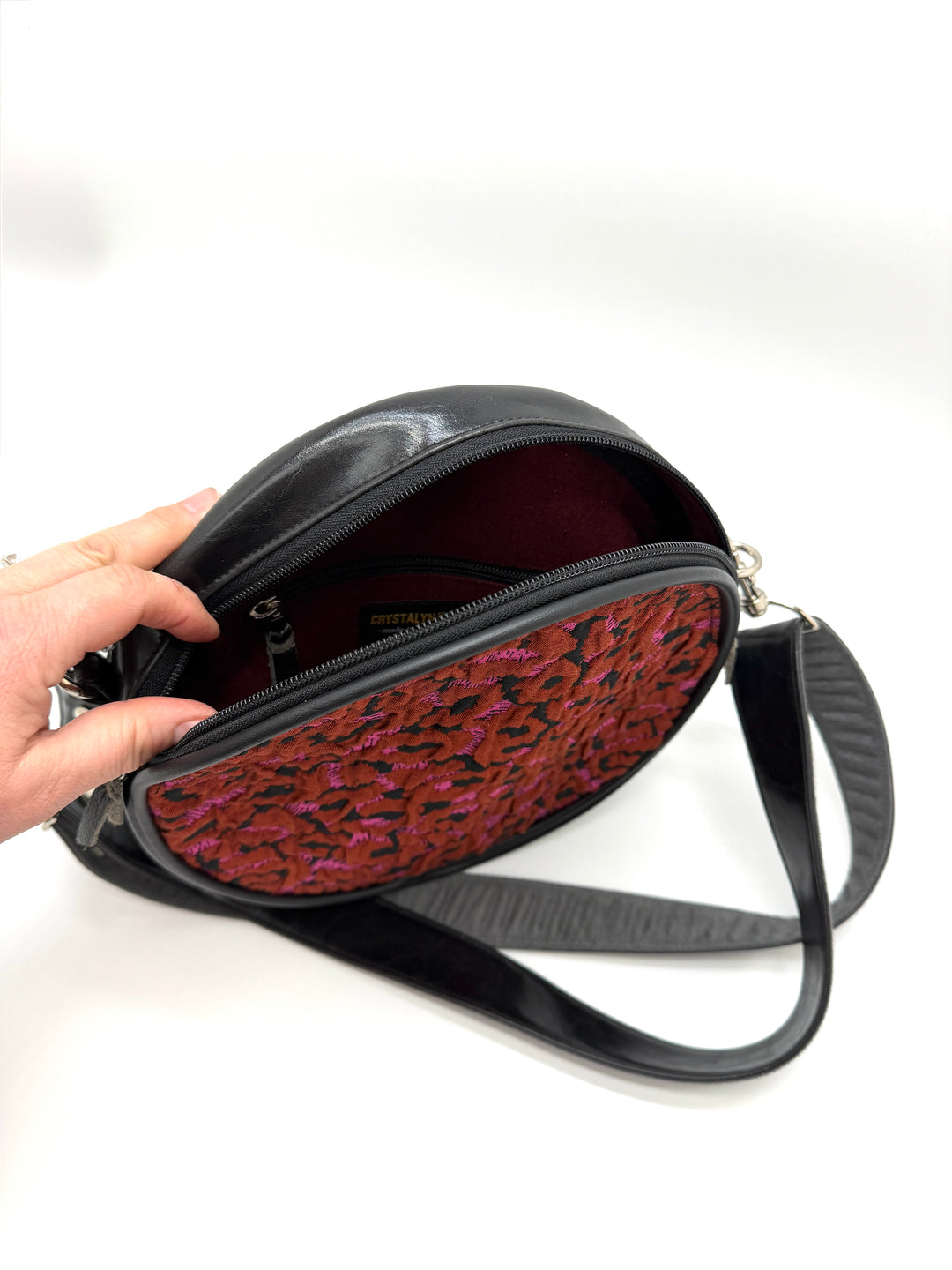 Black handbag with a red and black patterned interior, held open by a hand on a white background.#color_poppy-leopard