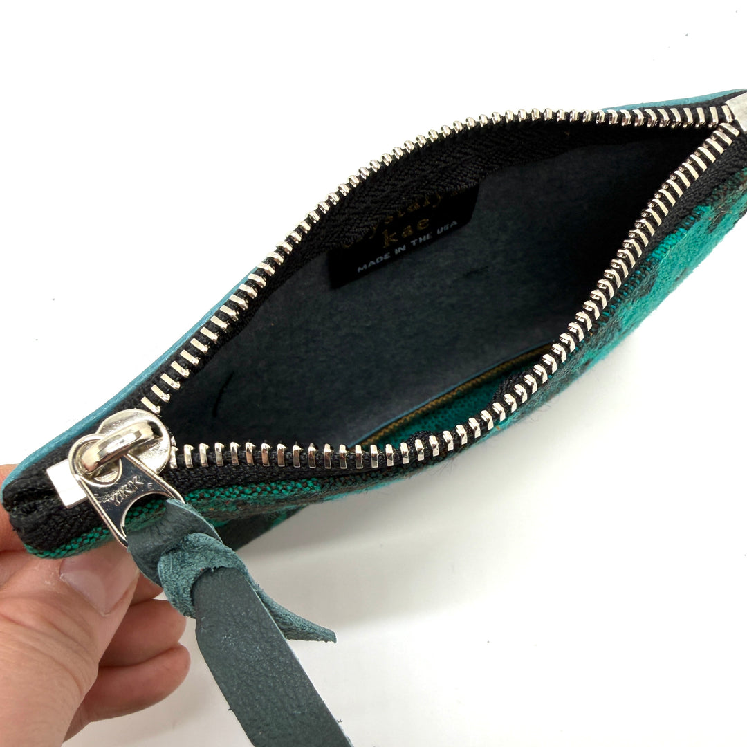 Teal clutch bag with a silver zipper held by a hand on a white background#color_teal-ferns