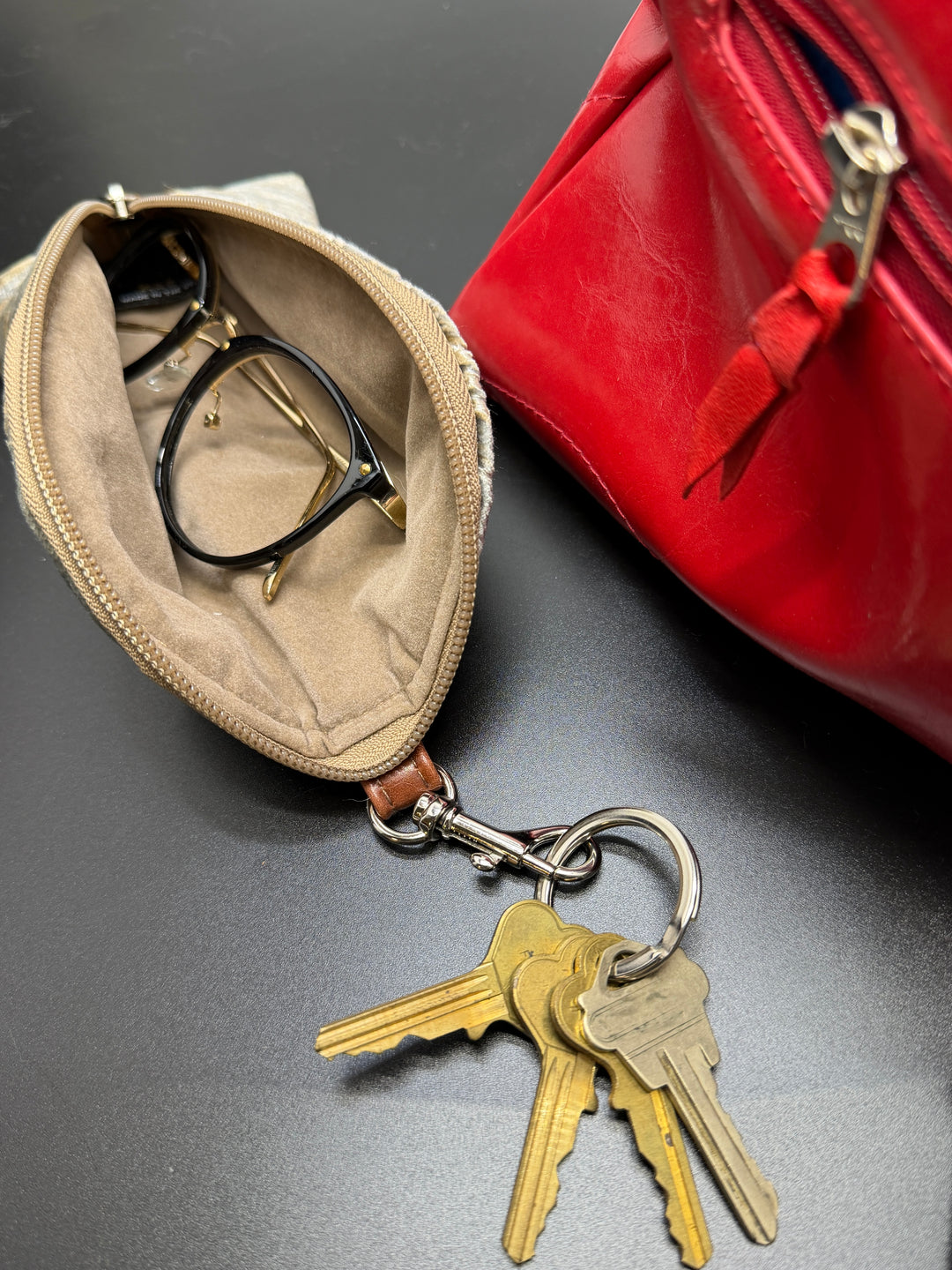 Keychain with keys attached to a beige pouch containing glasses, next to a red bag on a dark surface.