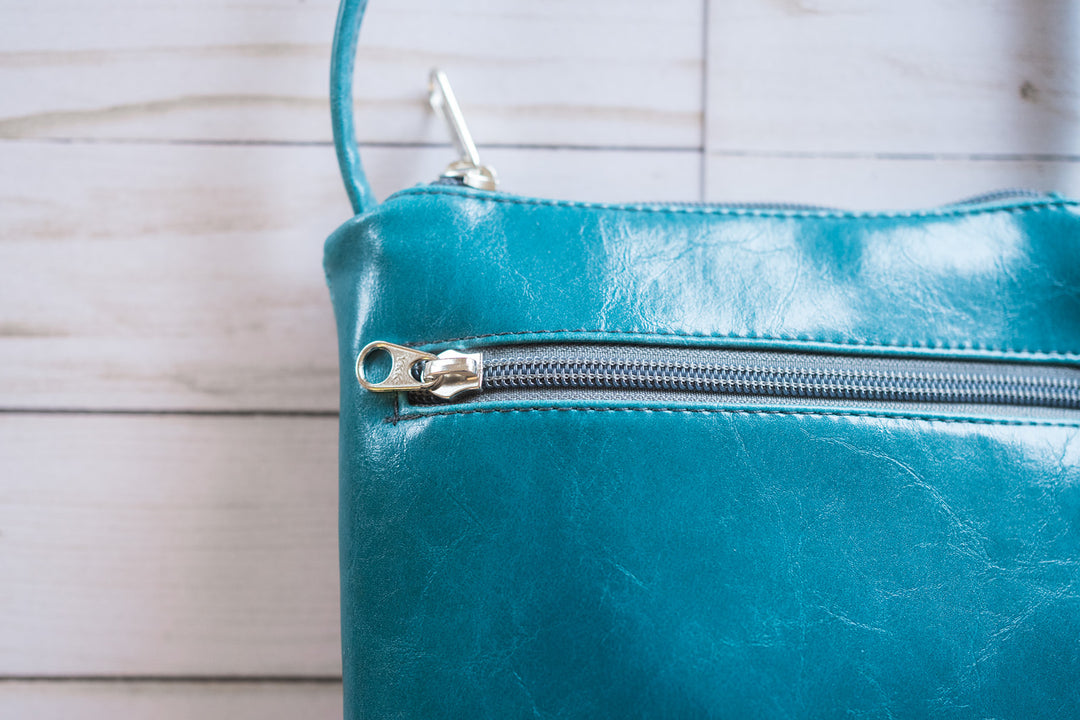 Teal leather clutch bag with a silver zipper on a wooden surface#color_arcade-dots-blue-green