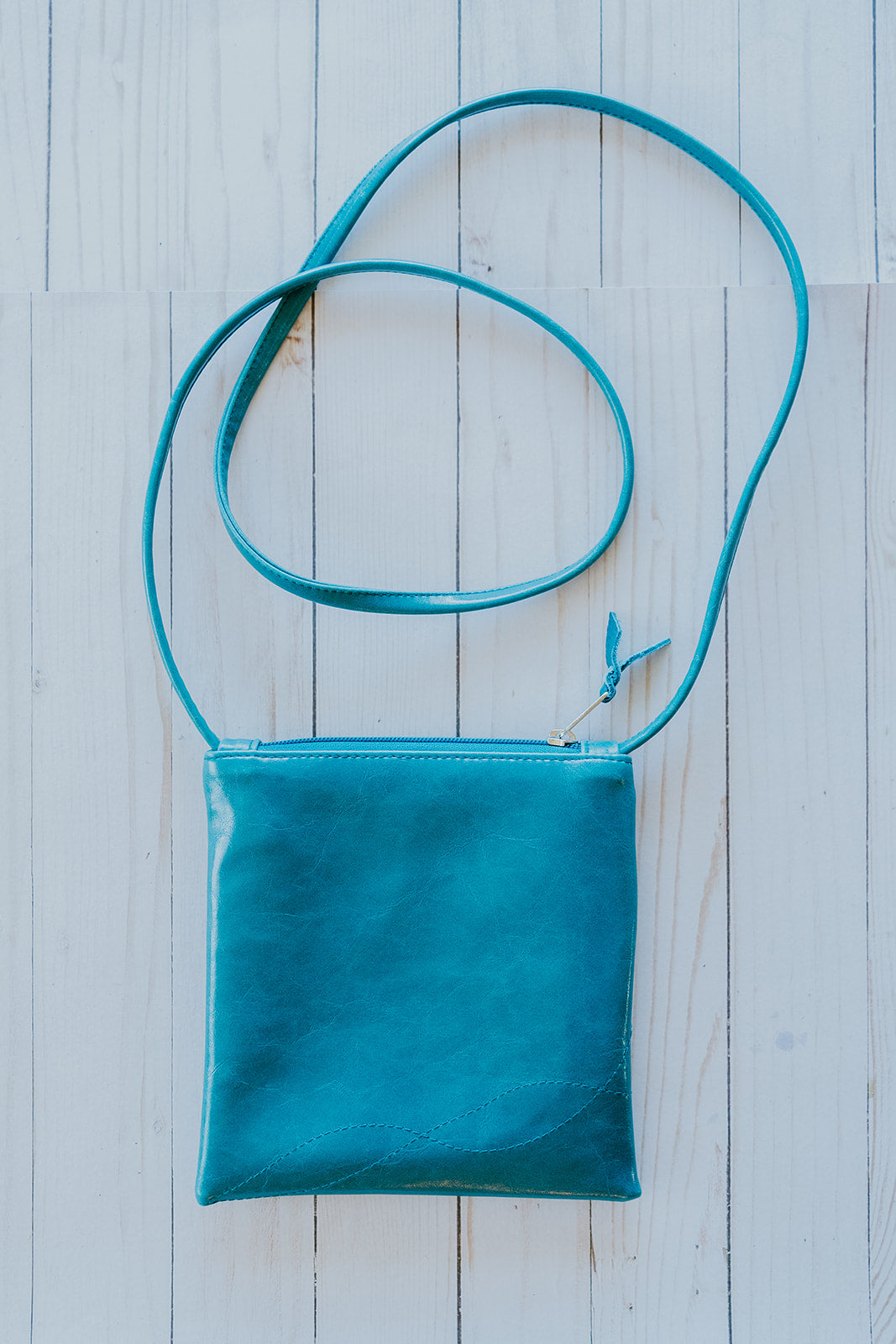 Teal leather crossbody bag on a light wooden surface#color_arcade-dots-blue-green