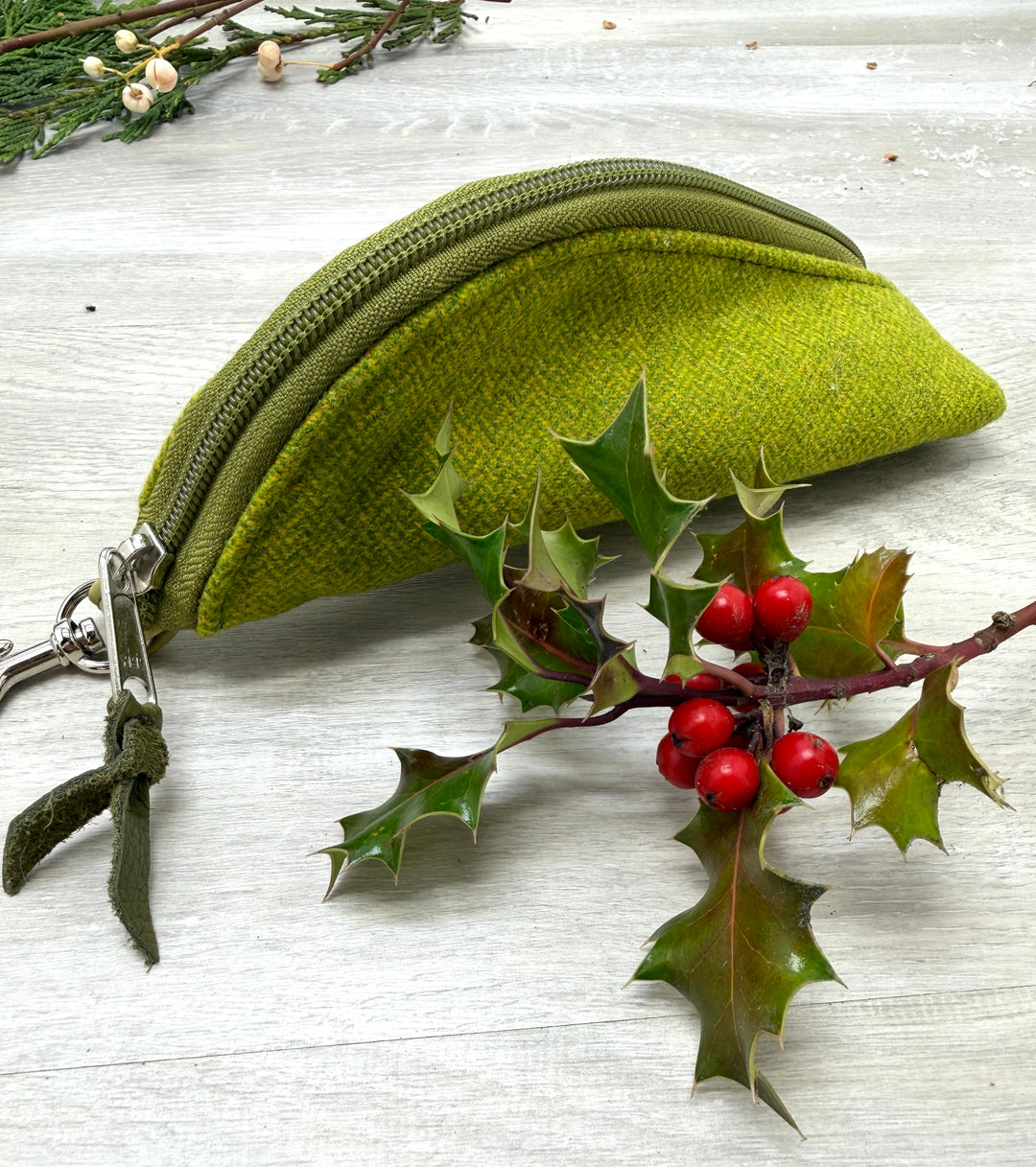 Green fabric pouch with a zipper on a light wooden surface with holly leaves and berries. #color_chartreuse-herringbone