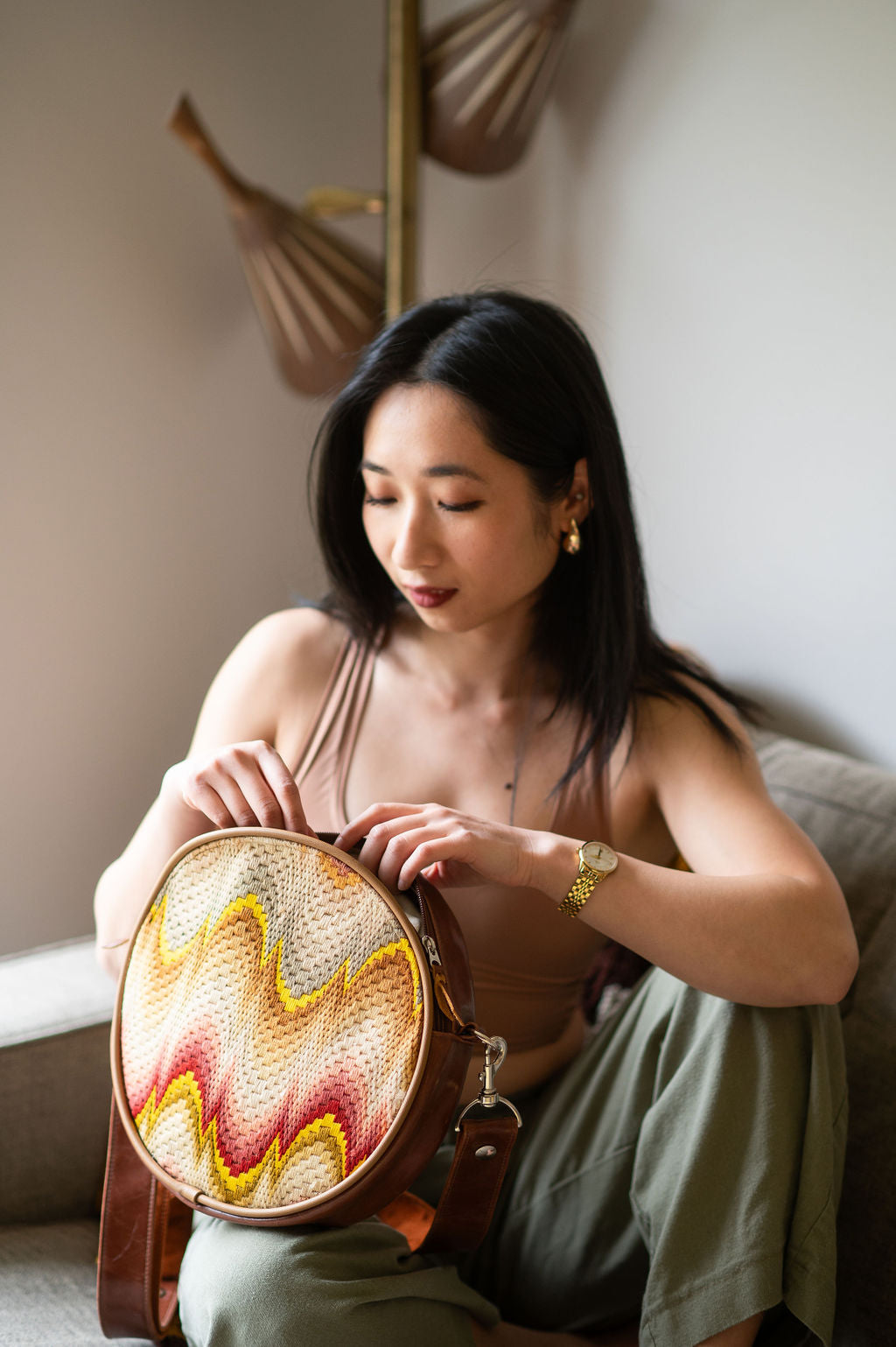 Woman holding a round, woven bag with a colorful pattern in a softly lit room.#color_bargello-needlepoint