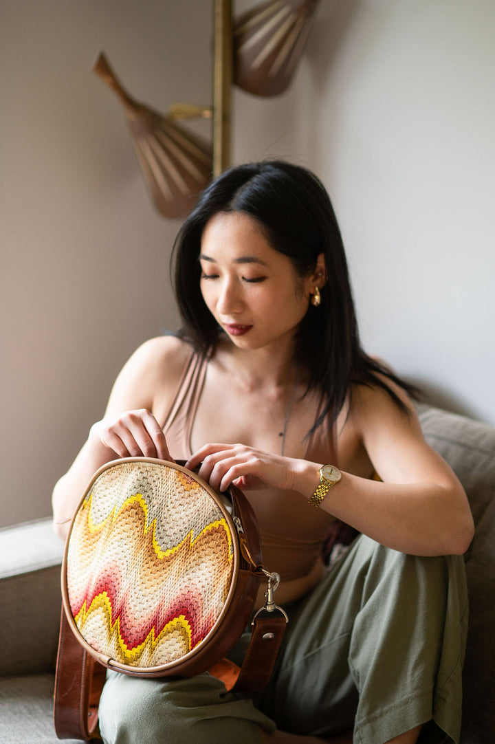 Woman holding a round, woven bag with a colorful pattern in a softly lit room.#color_bargello-needlepoint