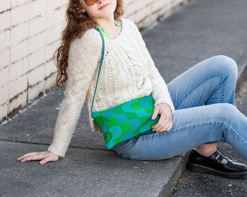 Woman sitting on a sidewalk holding a green purse, wearing sunglasses and a white sweater.