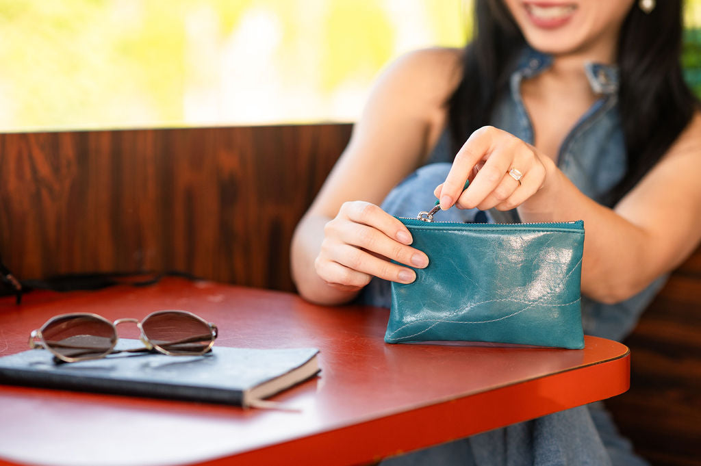 Woman opening a teal clutch at a table with sunglasses and a notebook.#color_teal