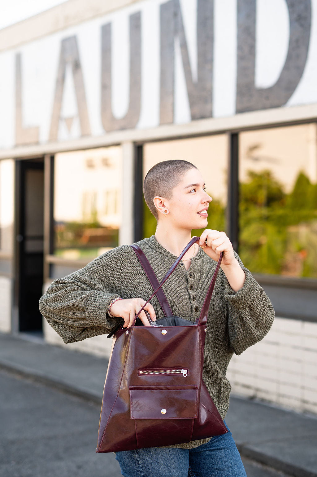 Person holding a brown leather backpack in front of a building with large letters that say laundromat. #color_wine