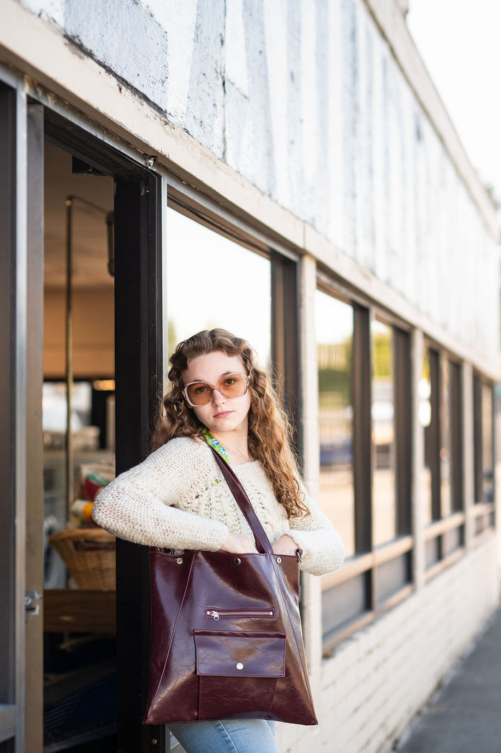 Woman holding a brown leather tote bag outdoors near a building with large windows. #color_wine