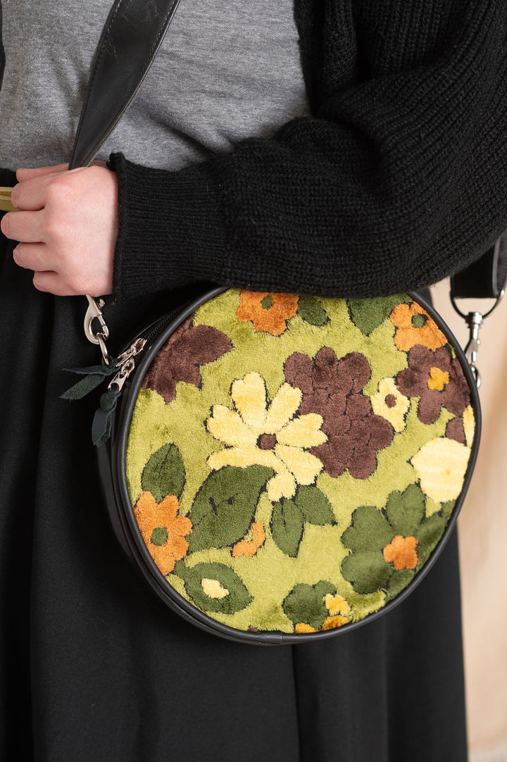 Round floral-patterned bag held by a person wearing a black sweater and gray shirt.#color_groovy-green-floral-chenille