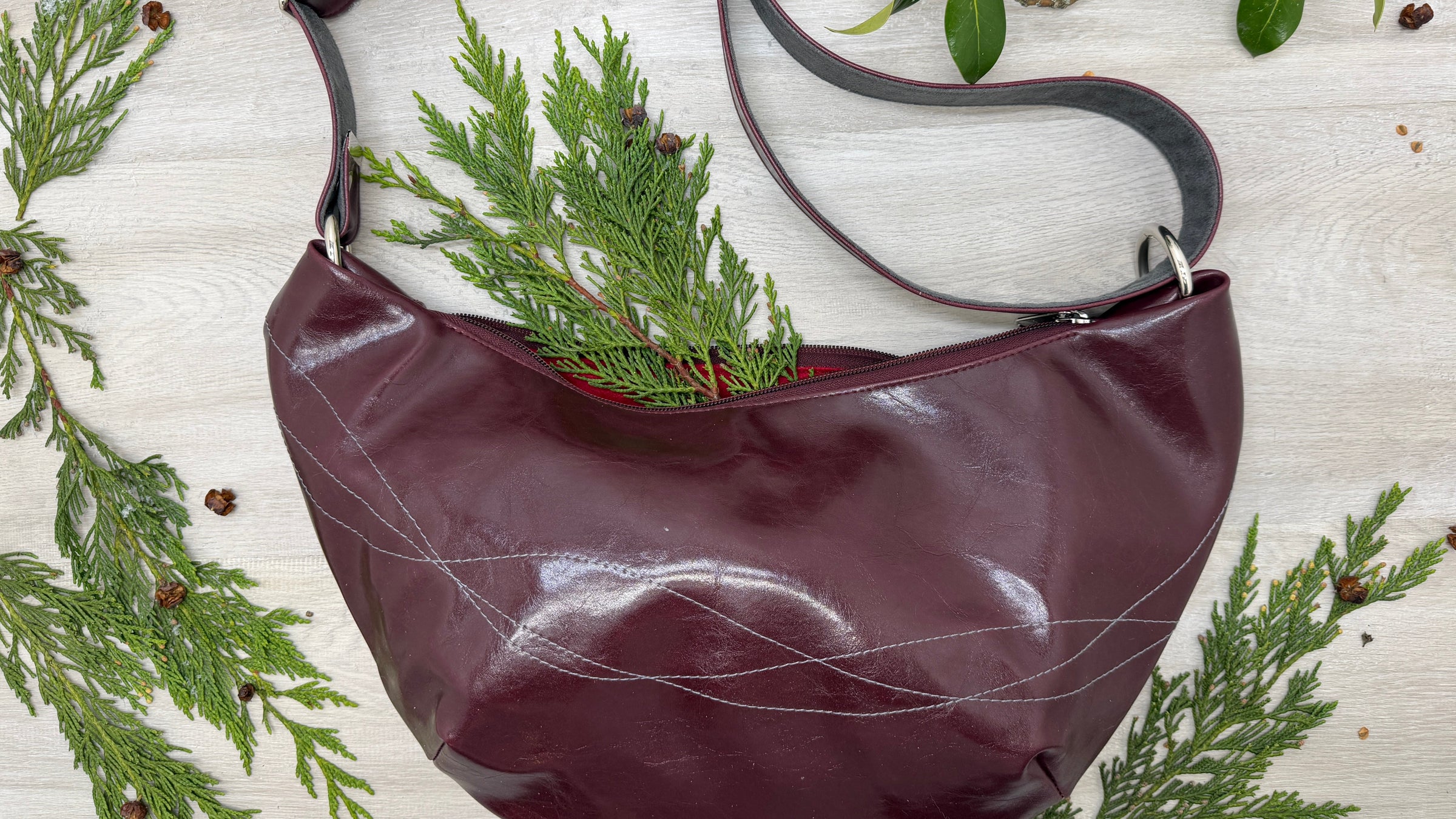 Burgundy wine vegan leather handbag with greenery on a light wooden surface with foliage.