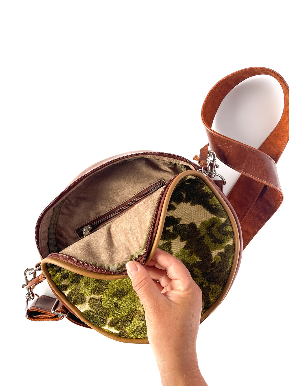 Hand holding a round green velvet patterned bag with a brown strap on a white background#color_ornate-mossy-green-chenille