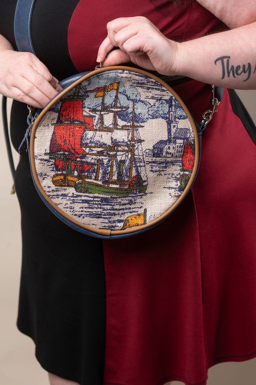 Person holding a round embroidered piece with a ship design, wearing a red dress.#color_nautical-sailboat-barkcloth